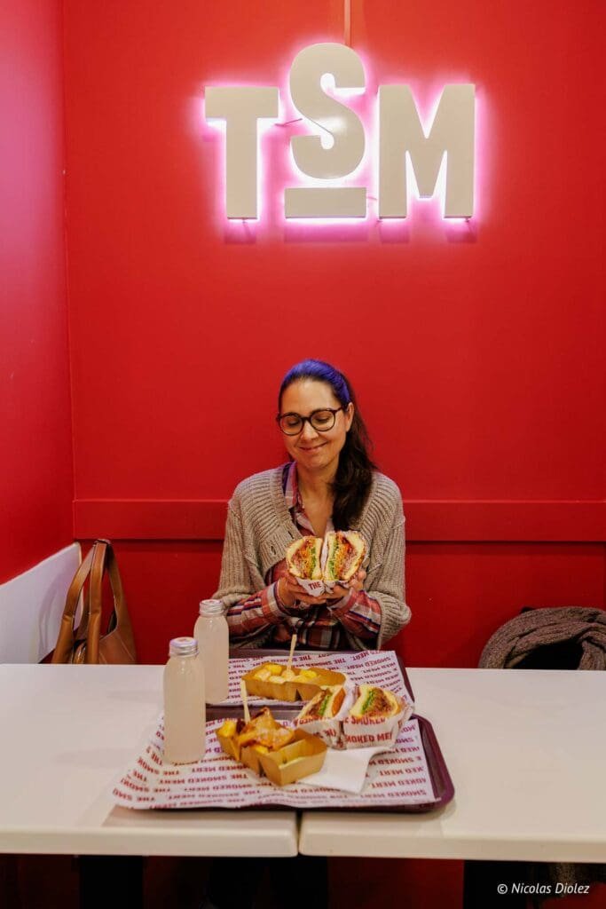 Femme souriante avec sandwich coupé devant le logo lumineux TSM rouge.