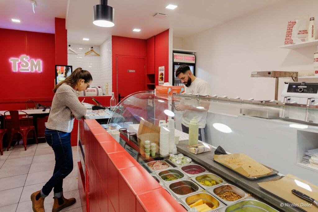 Client commande au comptoir de sandwicherie avec enseigne néon "TSM" rouge.