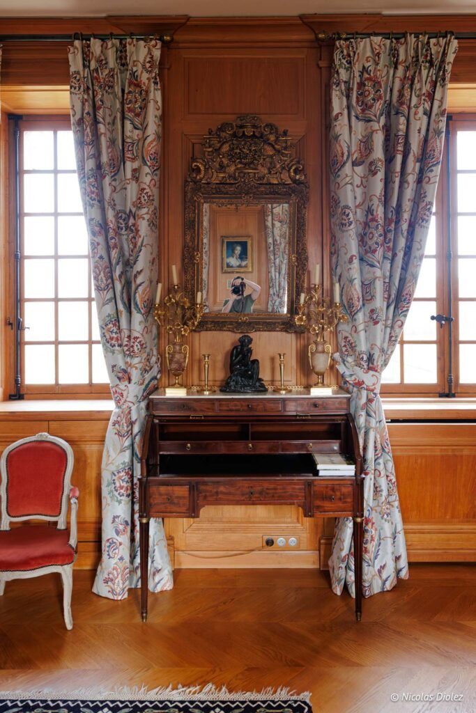 Bureau ancien en bois sous miroir doré, devant une fenêtre au Château de la Rivière.
