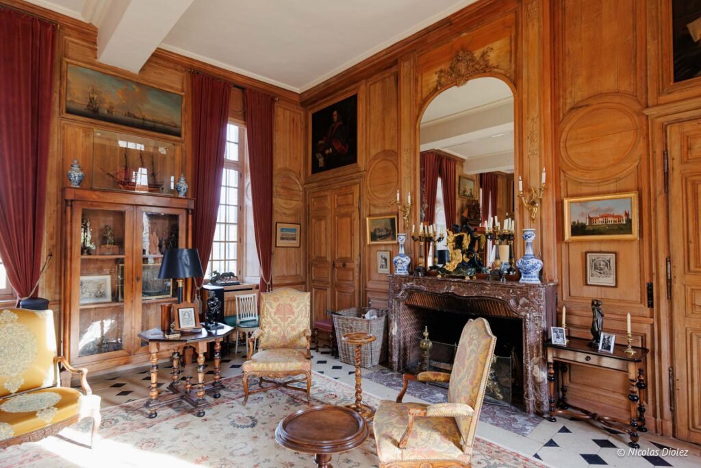 Salon boisé d'un château avec meubles anciens, cheminée marbre et miroir orné.
