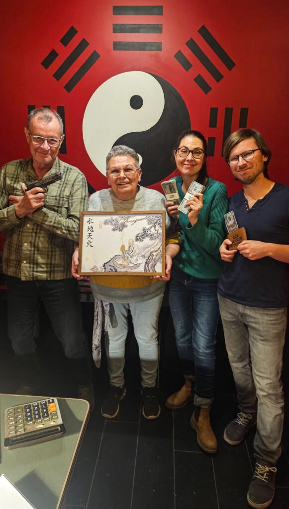 Quatre personnes posent devant un symbole yin-yang, tenant un tableau, des billets de banque et un pistolet.