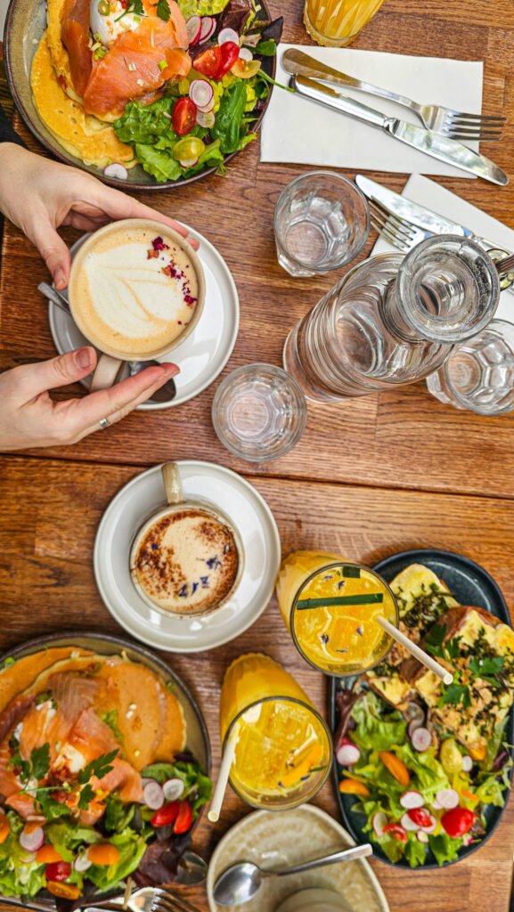 Brunch composé de pancakes au saumon fumé, boissons, café latte et salades fraîches sur une table en bois.