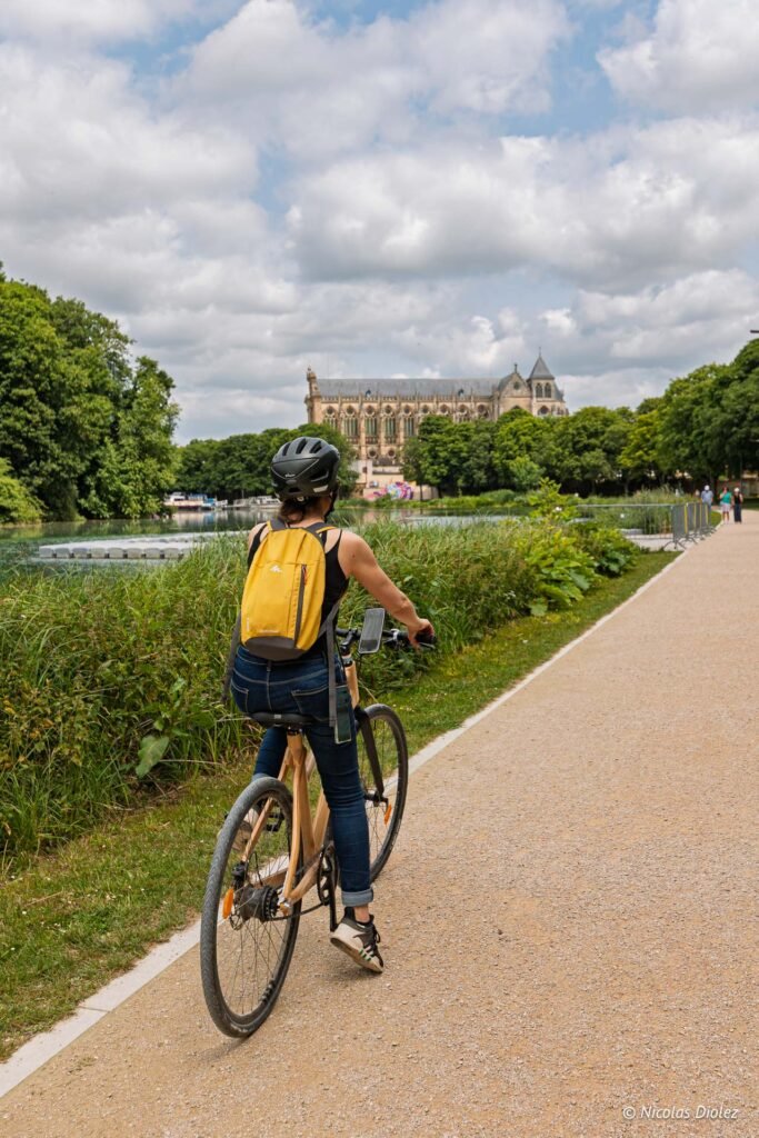 Cycliste avec sac à dos jaune sur une piste cyclable à Châlons-en-Champagne, avec la cathédrale en arrière-plan.