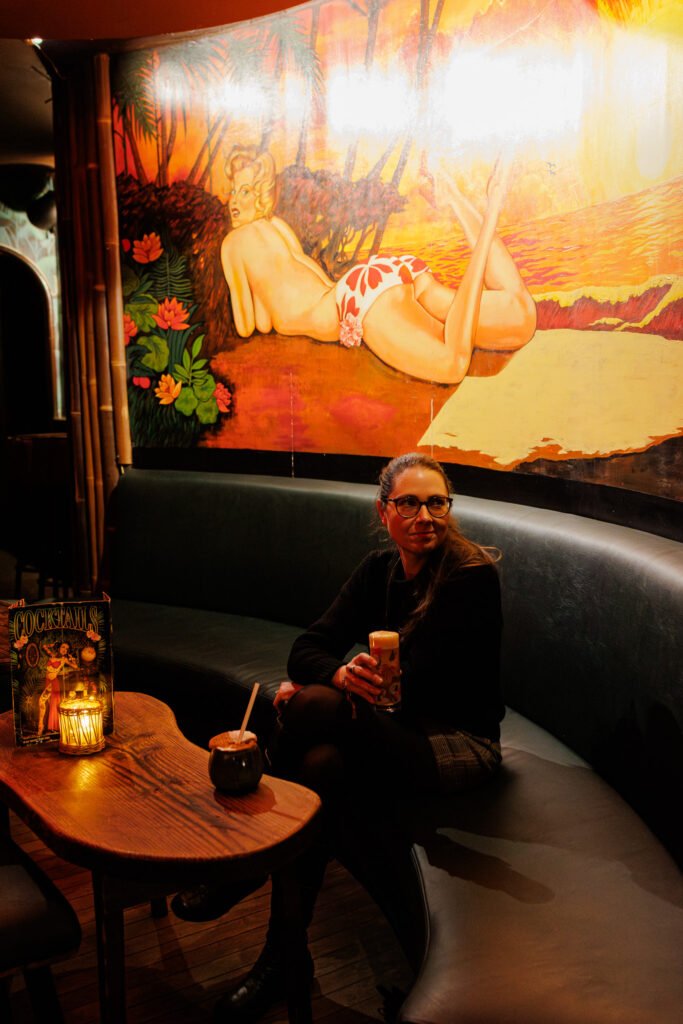 Femme tenant un cocktail dans un bar tiki à Paris devant une fresque murale pin-up vintage.