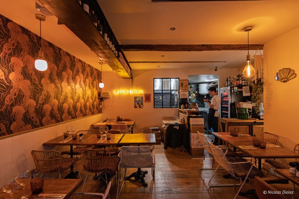 Intérieur chaleureux du restaurant La Prohibition à Sète avec tables, chaises et enseigne néon lumineuse.