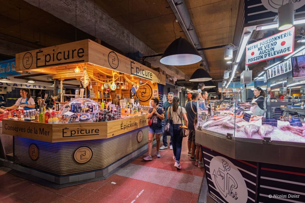 Clients dans les Halles de Sète devant l'épicerie fine Épicure et une poissonnerie locale.