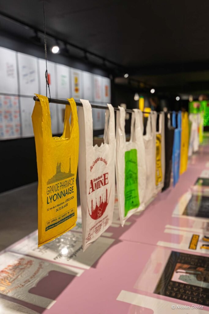 Sacs plastiques publicitaires suspendus lors d'une exposition au MIAM de Sète, avec logos de commerces lyonnais.