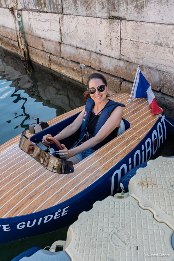 Femme souriante pilotant un Miniboat à Sète avec un drapeau français, près d'un quai en pierre.