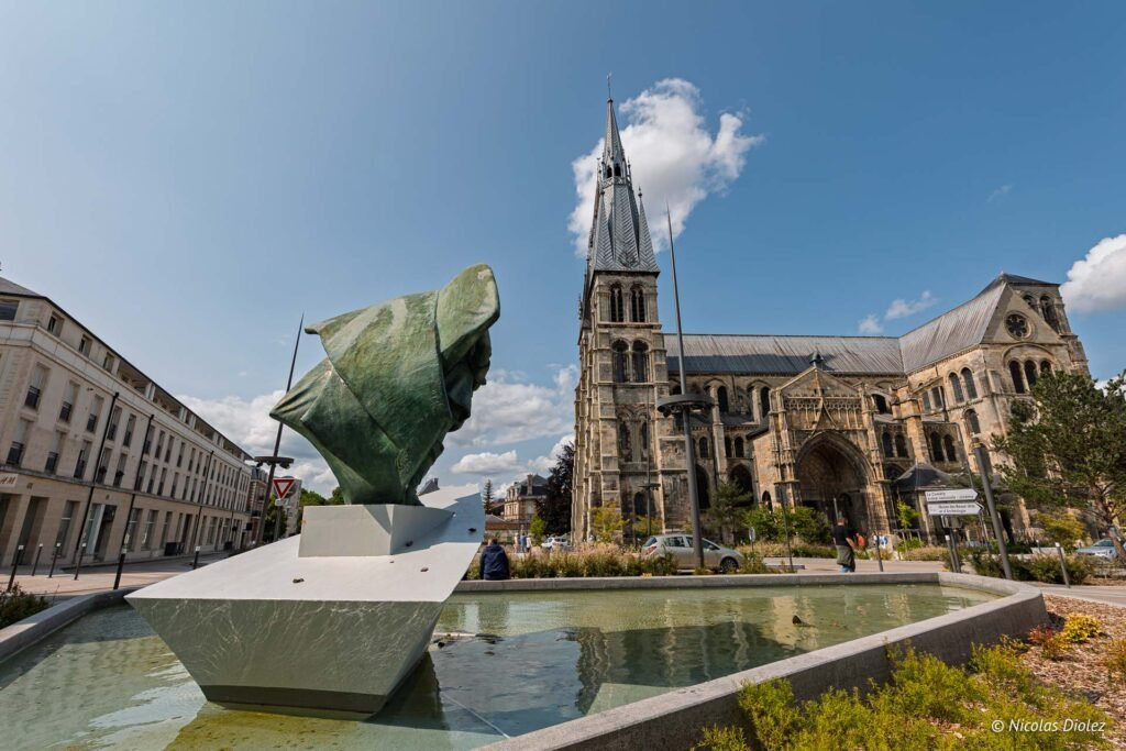 Sculpture contemporaine devant la collégiale Notre-Dame-en-Vaux à Châlons-en-Champagne sous un ciel bleu ensoleillé.