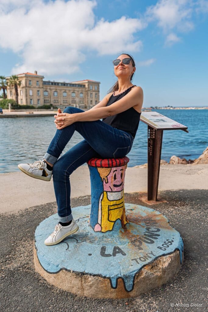 Femme assise sur une borne d'amarrage peinte à La Pointe Courte à Sète, près d'un panneau d'information touristique.