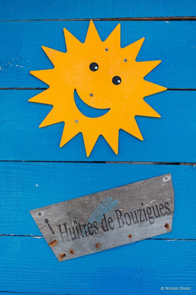 Panneau en bois Huîtres de Bouzigues avec un soleil souriant décoratif sur un mur bleu à La Pointe Courte.