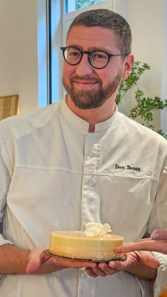 Le chef pâtissier Dan Touati présentant un entremets élégant décoré d'une fleur en chocolat blanc.