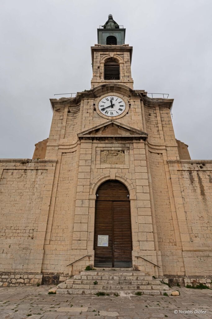 Façade en pierre de l'église Saint-Louis à Sète avec son clocher, son horloge et son grand portail en bois.