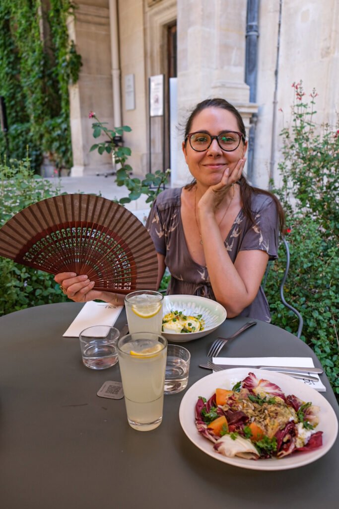 Femme souriante avec un éventail à une table de restaurant, accompagnée de limonades et de salades.