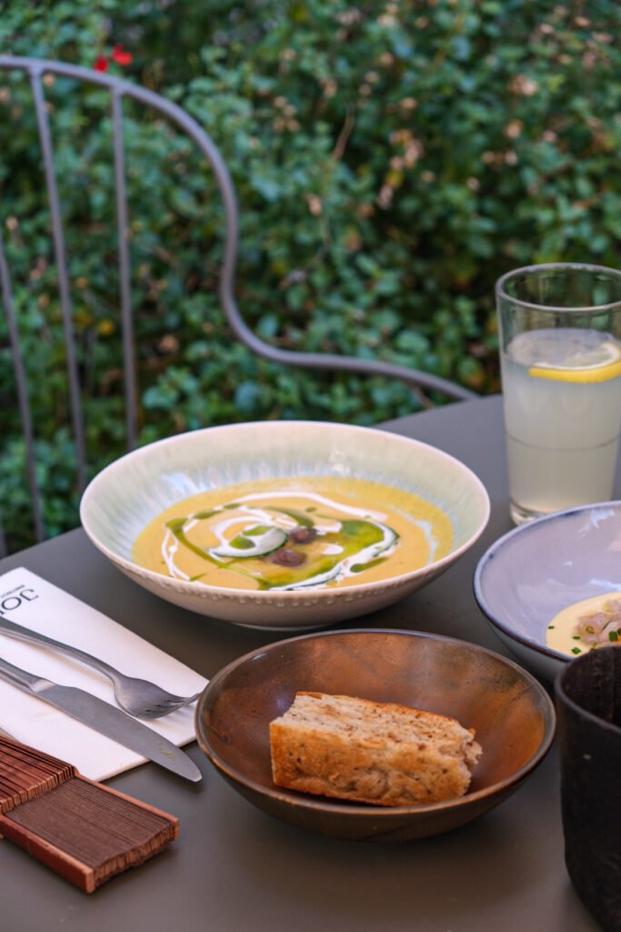 Bol de soupe crémeuse avec huile aux herbes et crème, accompagné de pain, sur une terrasse en extérieur.