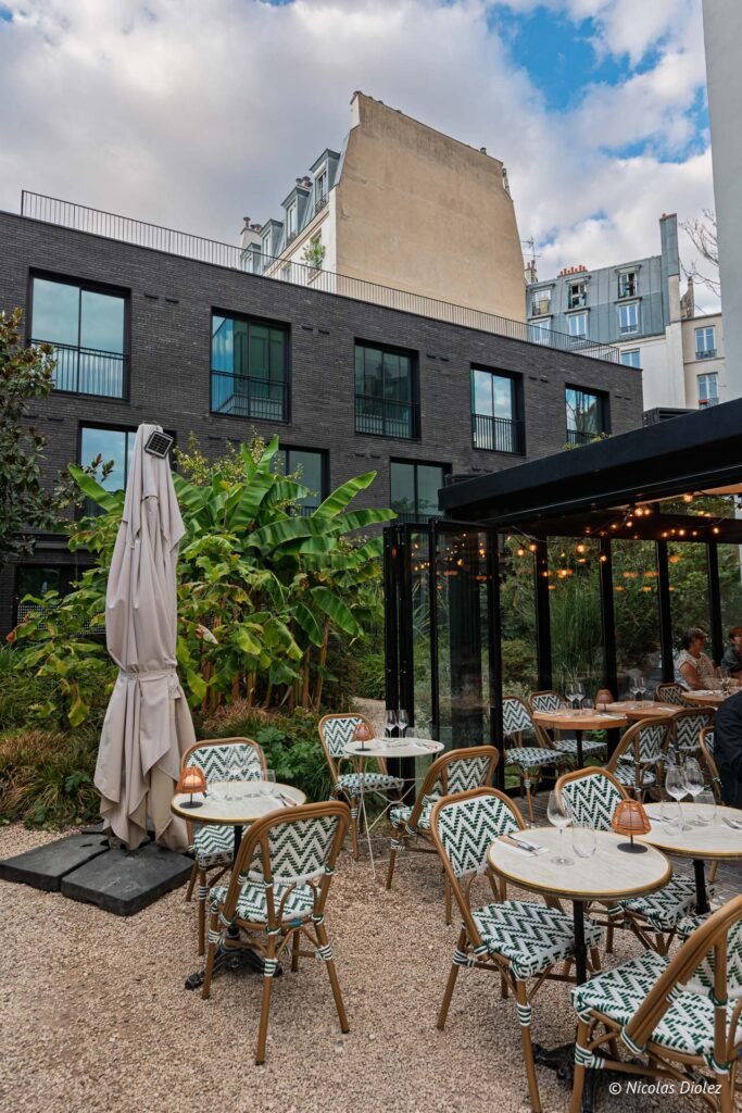 Terrasse du restaurant Laia à Paris avec mobilier de bistrot, nichée dans un jardin urbain avec bâtiment moderne.