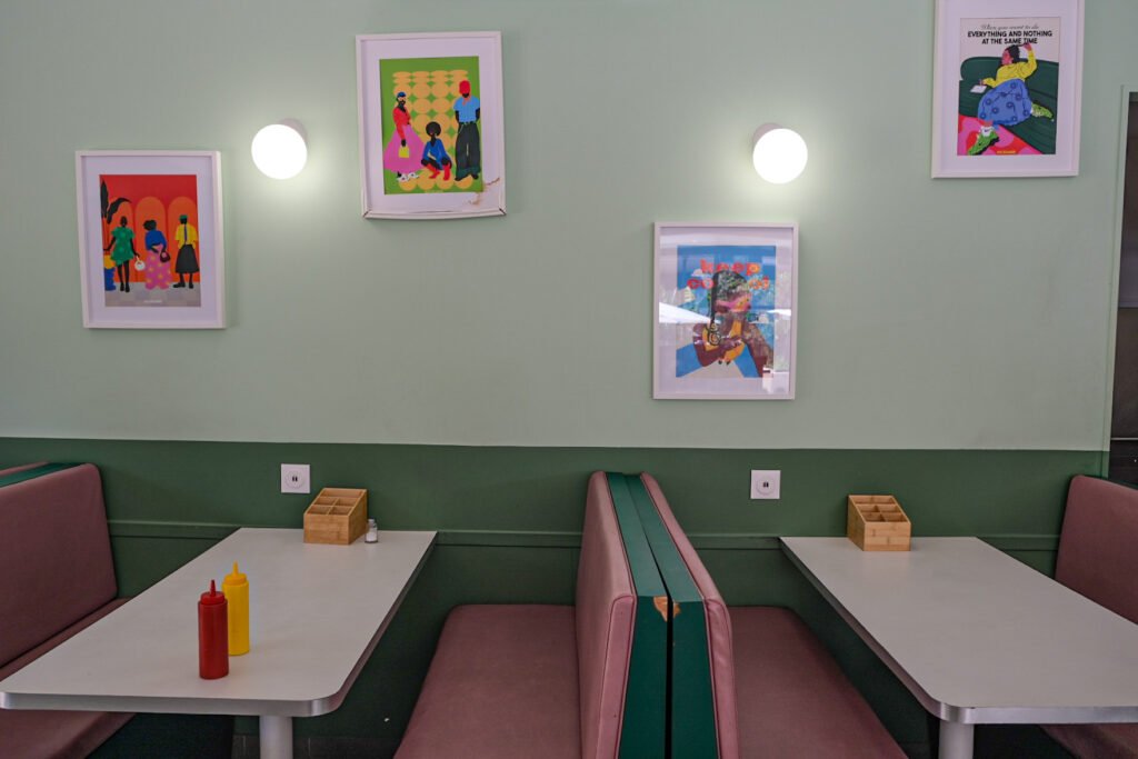 Tables et banquettes roses dans un restaurant décoré d'affiches illustrées colorées sur un mur vert.