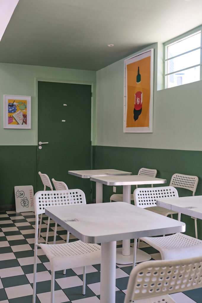Intérieur de café avec tables blanches, chaises perforées, sol en damier et panneaux décoratifs muraux.