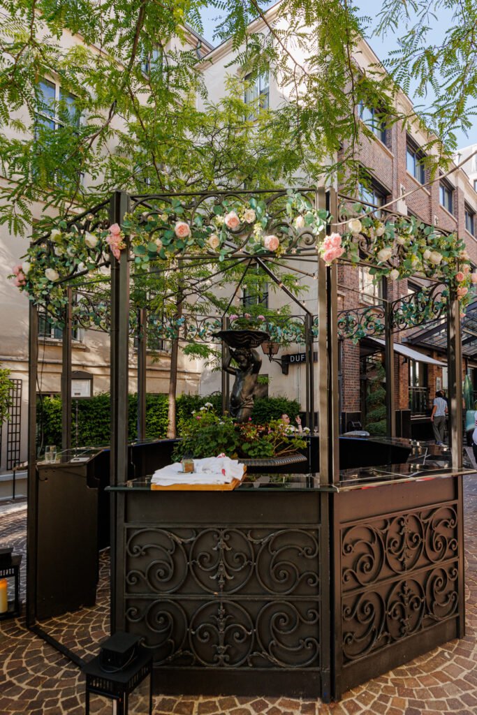 Bar extérieur en fer forgé avec fontaine décorative au cœur des Jardins du Marais à Paris.