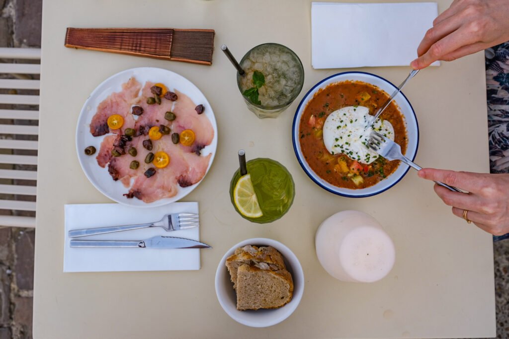 Repas estival composé de carpaccio de poisson, burrata sur légumes, boissons fraîches et pain sur une table.