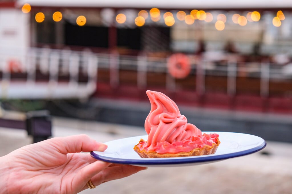 Main tenant une assiette avec une tartelette aux fruits surmontée d'une crème fouettée rose à bord d'un bateau.