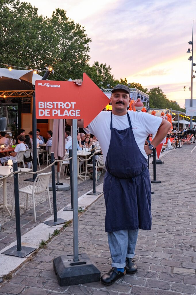 Serveur portant un tablier à côté d'un panneau indicateur rouge Plat-Form Bistrot de Plage devant une terrasse animée.