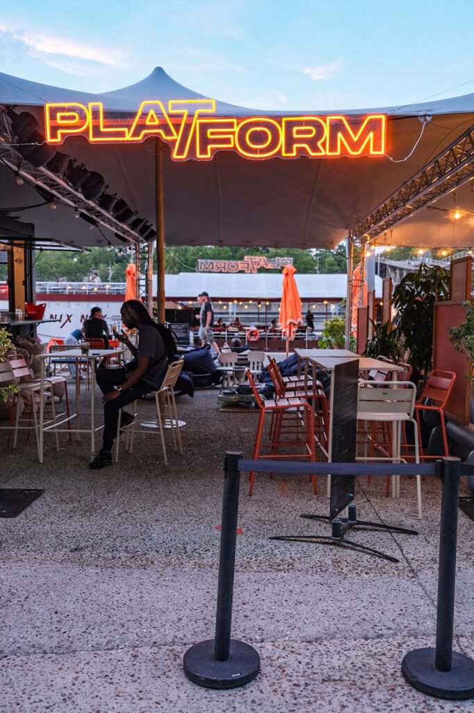 Terrasse du bar Plat-Form en bord de Seine avec tables, chaises et enseigne néon lumineuse.