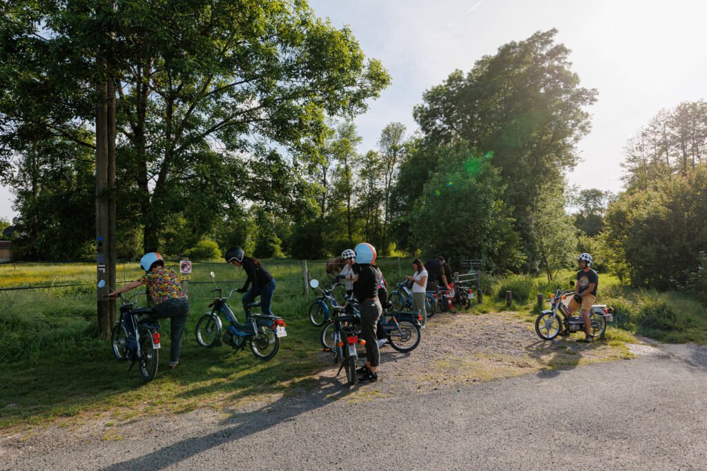 Groupe de personnes en mobylettes faisant une pause sur un chemin en bordure de forêt.