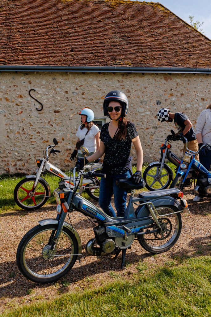 Femme en casque et lunettes avec sa mobylette Motobécane 51 devant un mur en pierre, entourée d'autres motards.