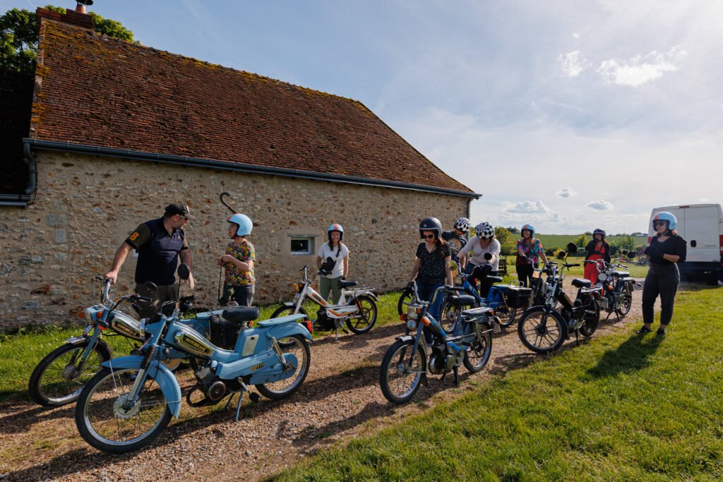 Groupe de personnes avec des mobylettes anciennes devant une bâtisse en pierre lors d'une balade Les Mobylettes de Marinette.