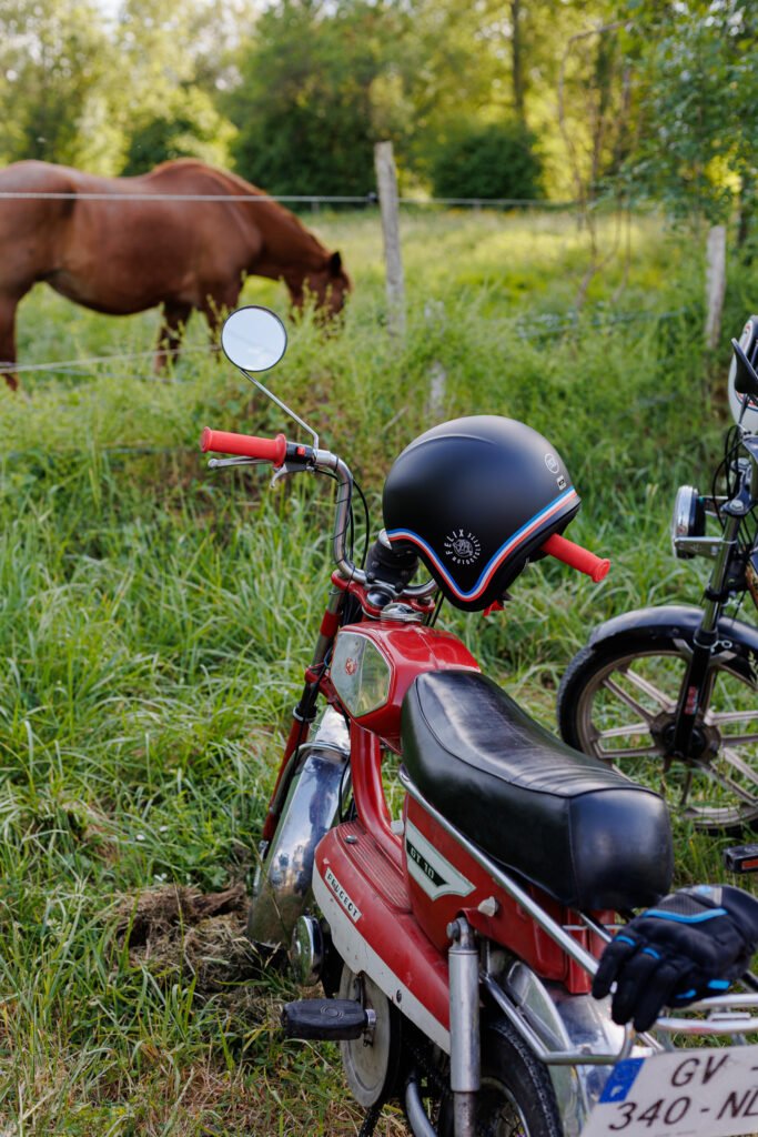 Mobylette Peugeot GT 10 rouge avec casque Felix Moto posé sur le guidon dans un champ avec un cheval.