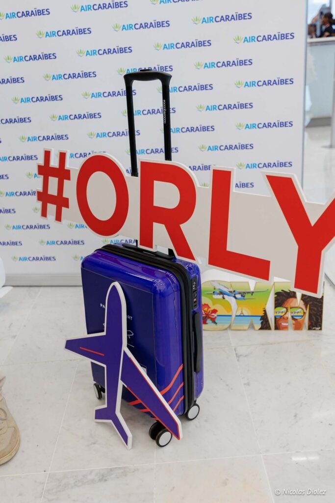 Valise Air Caraïbes devant un panneau de vol inaugural avec l'inscription Orly.