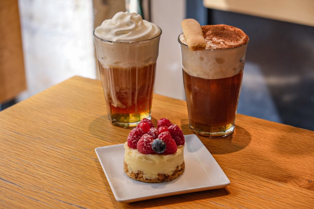 Two gourmet coffees with whipped cream and biscuits accompanied by a raspberry tartlet on a wooden table.