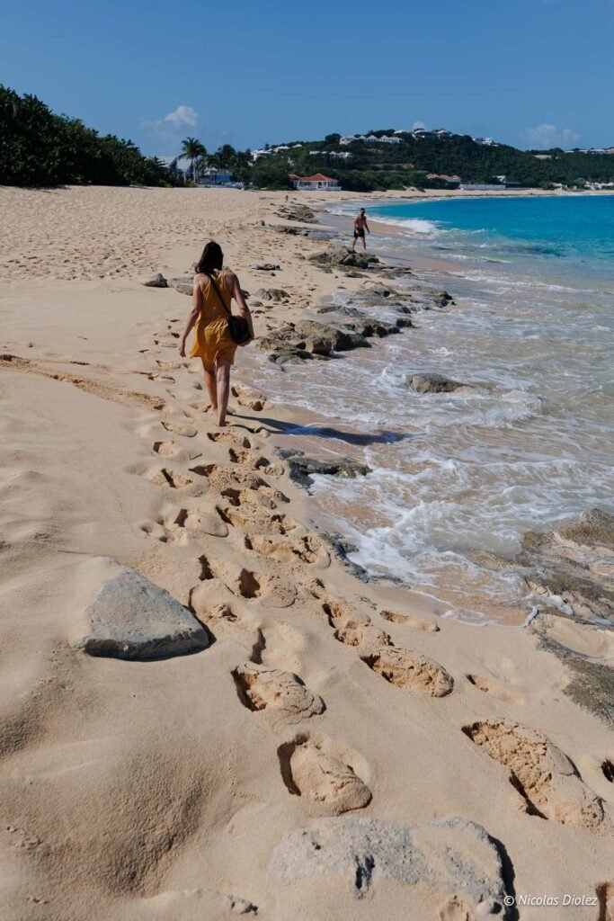 Femme marchant sur le sable de la plage de Baie Rouge à Saint-Martin avec des vagues et des collines en arrière-plan.