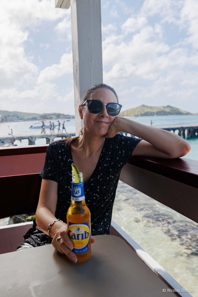 Femme souriante avec des lunettes de soleil tenant une bouteille de bière Carib dans un restaurant à Grand-Case, Saint-Martin.