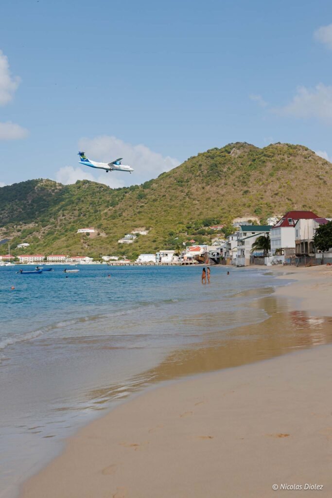 Avion atterrissant au-dessus de la plage de Grand-Case à Saint-Martin avec des collines verdoyantes en arrière-plan.