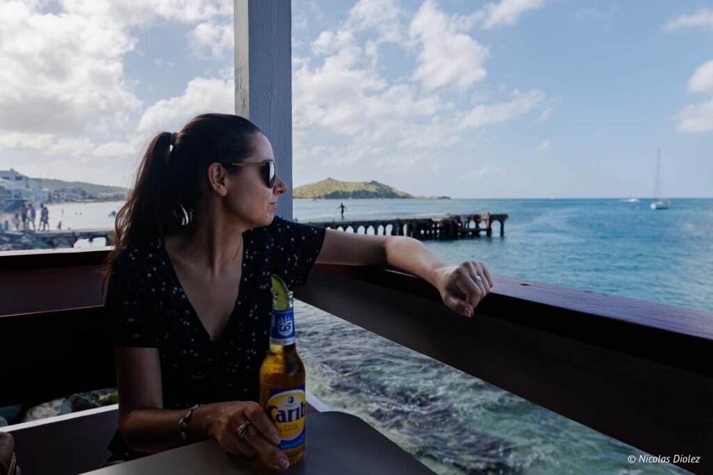 Femme assise à une terrasse de Grand-Case à Saint-Martin, tenant une bière Carib face à la mer.