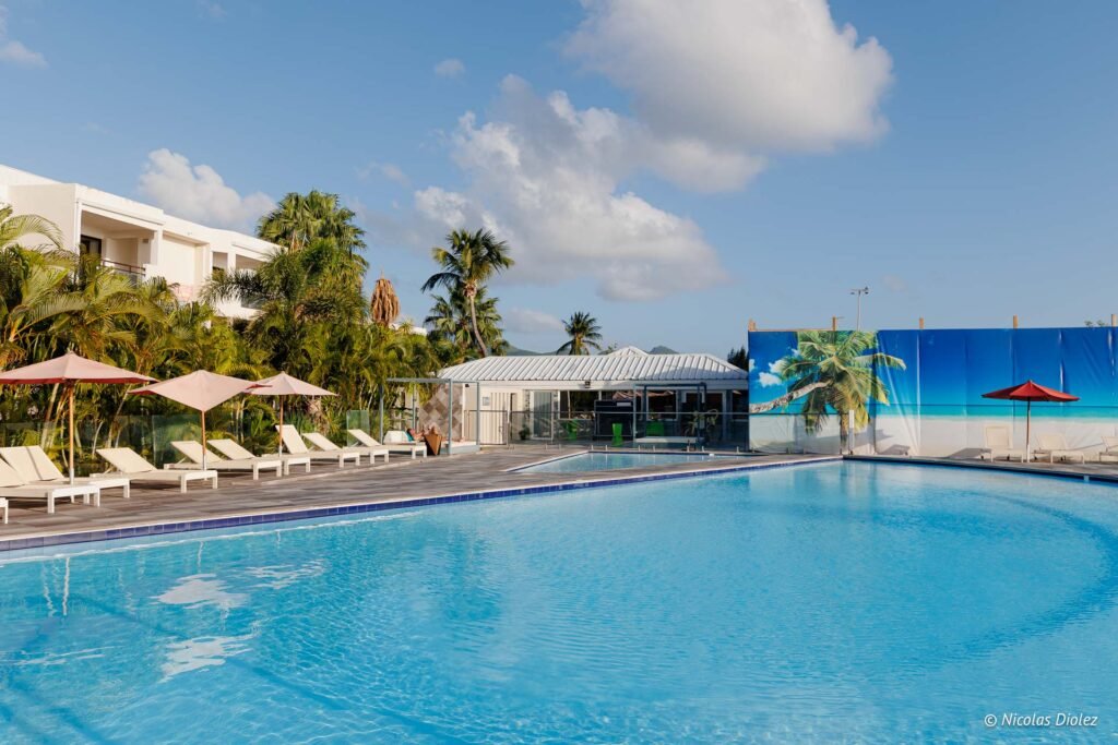 Piscine extérieure et transats de l'hôtel Hommage à Saint-Martin, entourée de palmiers sous un ciel ensoleillé.