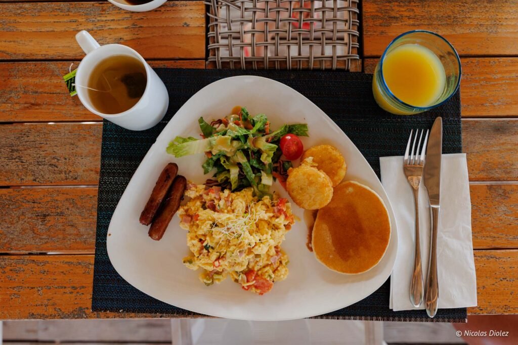 Assiette de petit-déjeuner salé comprenant des œufs brouillés, des saucisses, des pancakes, une salade et un jus de fruits.