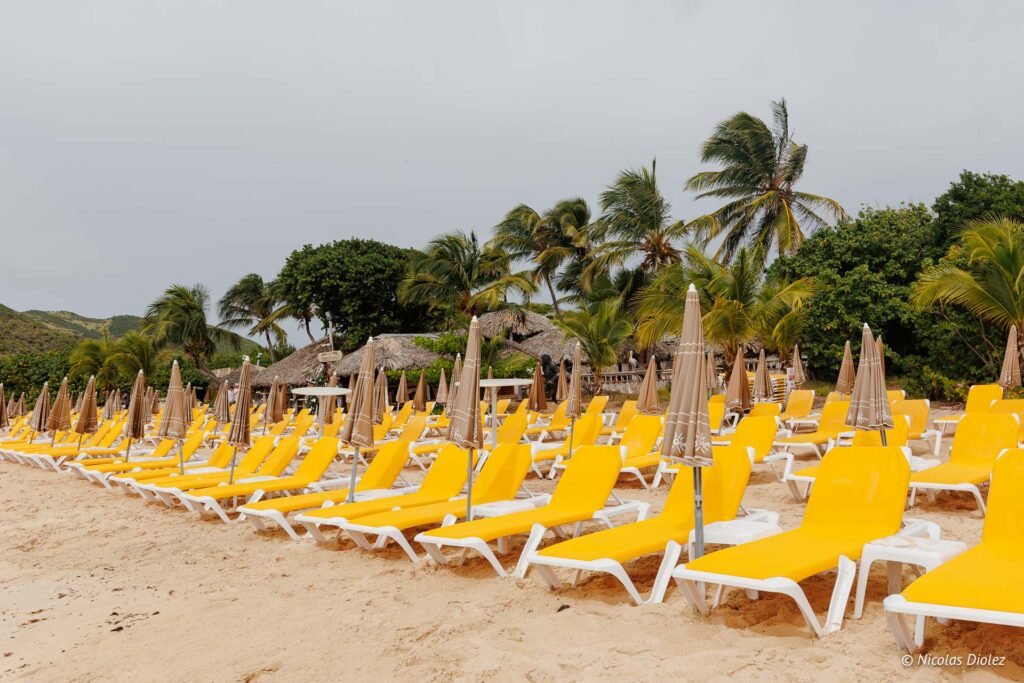 Transats jaunes et parasols fermés alignés sur une plage de sable de l'îlet Pinel sous un ciel nuageux.