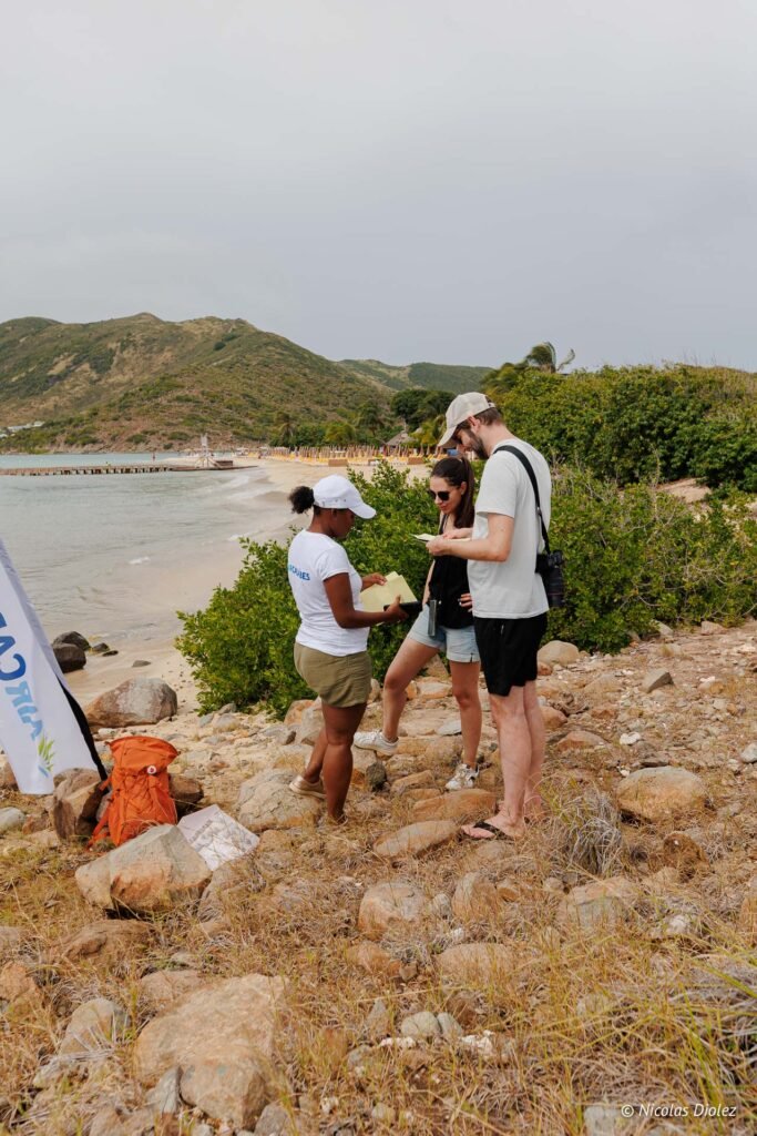 Trois personnes analysent des documents sur la plage de l'îlet Pinel sous un ciel nuageux.