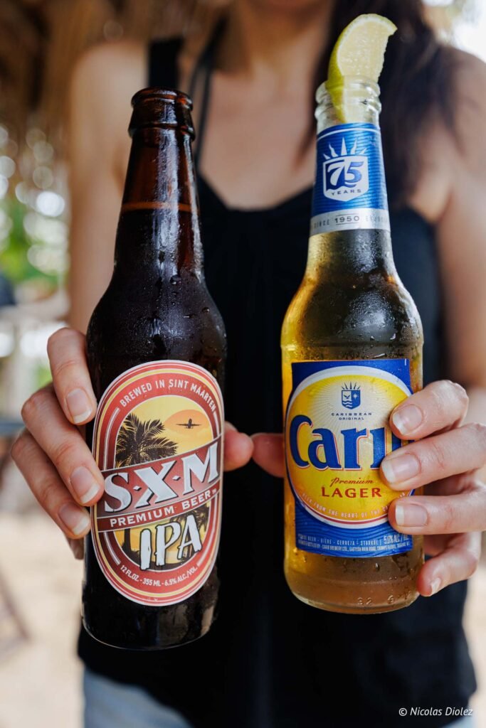 Une personne tenant des bouteilles de bière SXM IPA et Carib à l'Îlet Pinel.