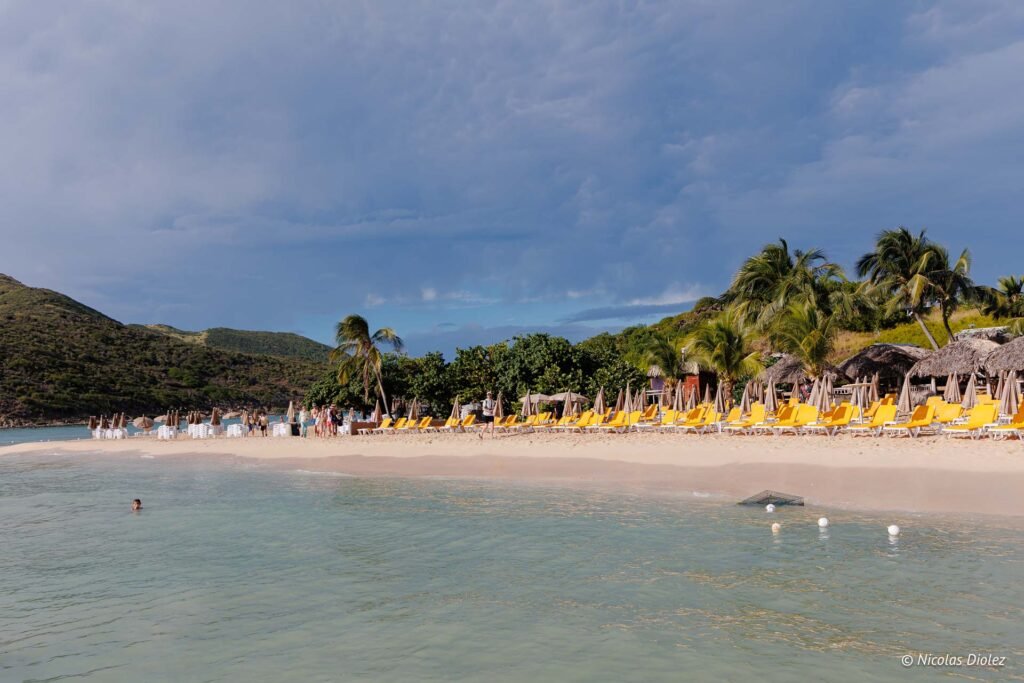 Plage paradisiaque de l'Îlet Pinel avec ses transats jaunes, ses palmiers et son eau turquoise sous un ciel nuageux.