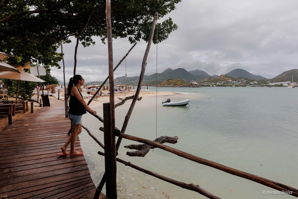 Femme sur une terrasse en bois surplombant la plage de l'îlet Pinel sous un ciel nuageux.