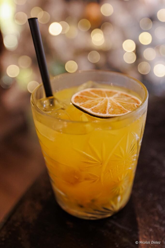 Cocktail rafraîchissant garni d'une tranche de citron déshydratée et d'une paille noire, servi dans un verre ciselé.