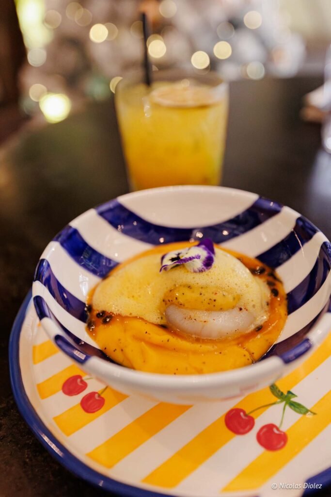 Plat gastronomique à base de Saint-Jacques servi dans un bol rayé avec un cocktail en arrière-plan chez Le Temps des Cerises.