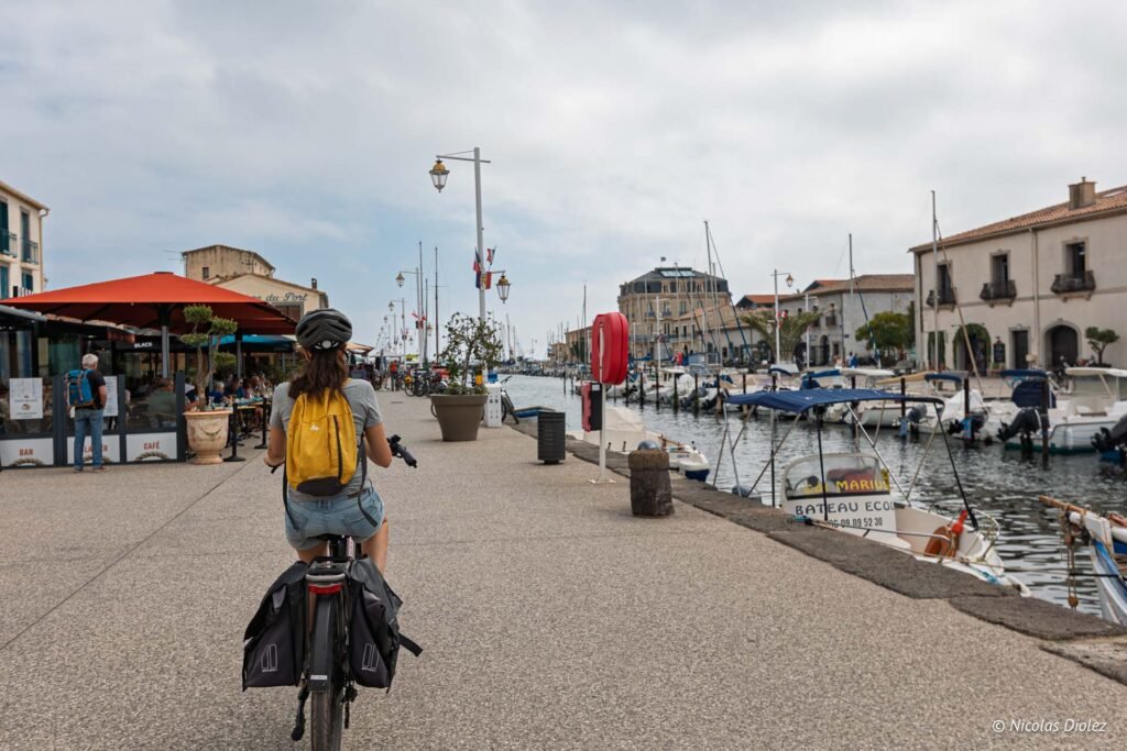 Cycliste à vélo sur le quai du port de Marseillan avec ses bateaux et ses terrasses de café.