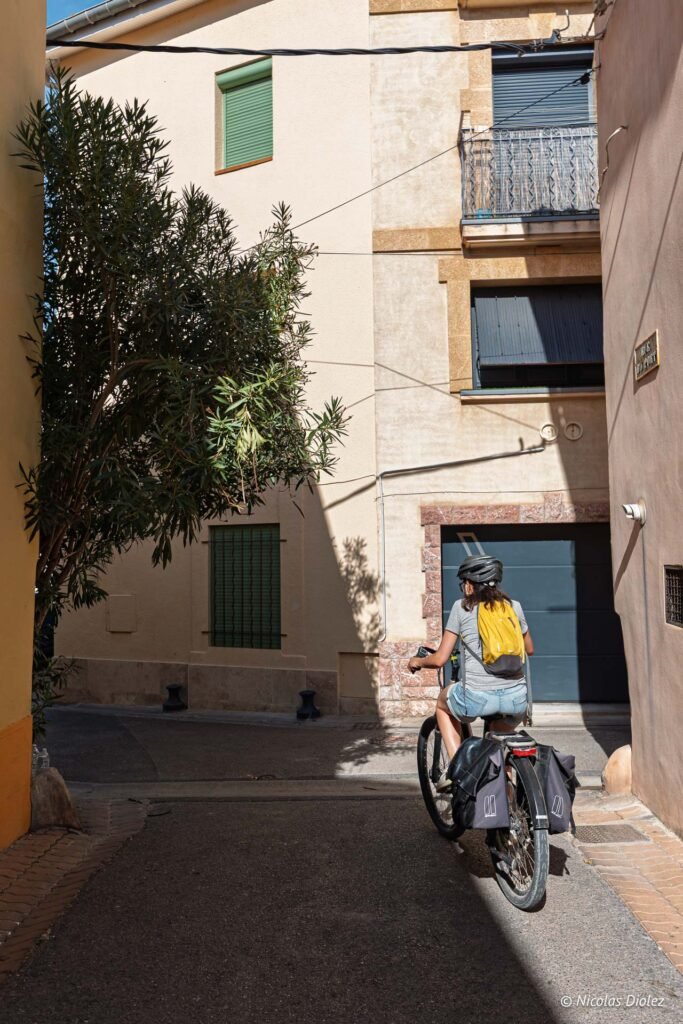 Cycliste avec sac à dos jaune à vélo dans une ruelle étroite et ensoleillée de Mèze.