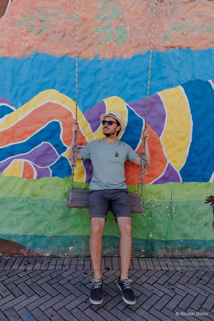 Homme souriant avec chapeau et lunettes de soleil se balançant devant un mur coloré à Saint-Martin.
