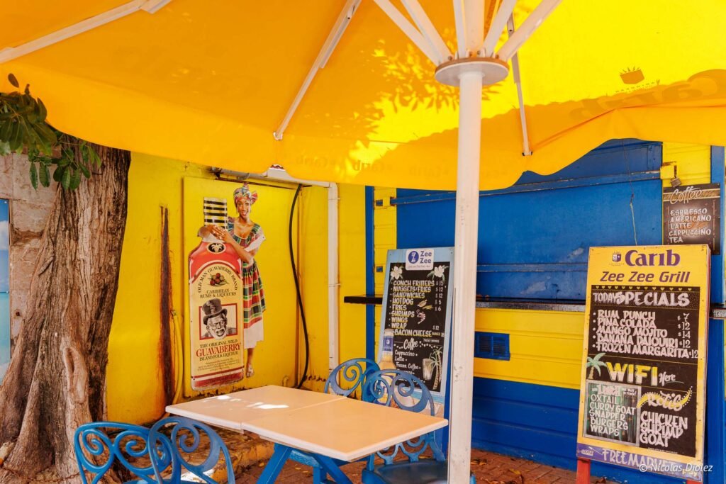 Terrasse ombragée d'un restaurant coloré en jaune et bleu avec des tables et un tableau des menus à Saint-Martin.